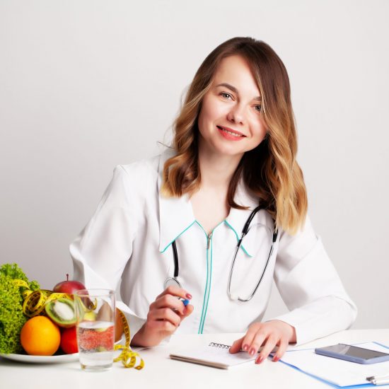 Young dietitian doctor at the consulting room at the table with fresh vegetables and fruits, working on a diet plan.